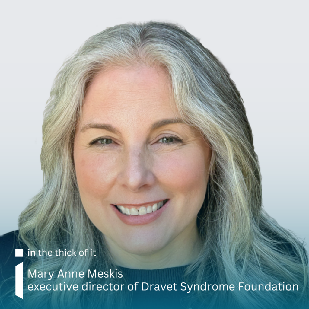 Image of Mary Anne Meskis looking into the camera and smiling. She has long grey/blond wavy hair and is wearing a dark jumper. The background is grey.