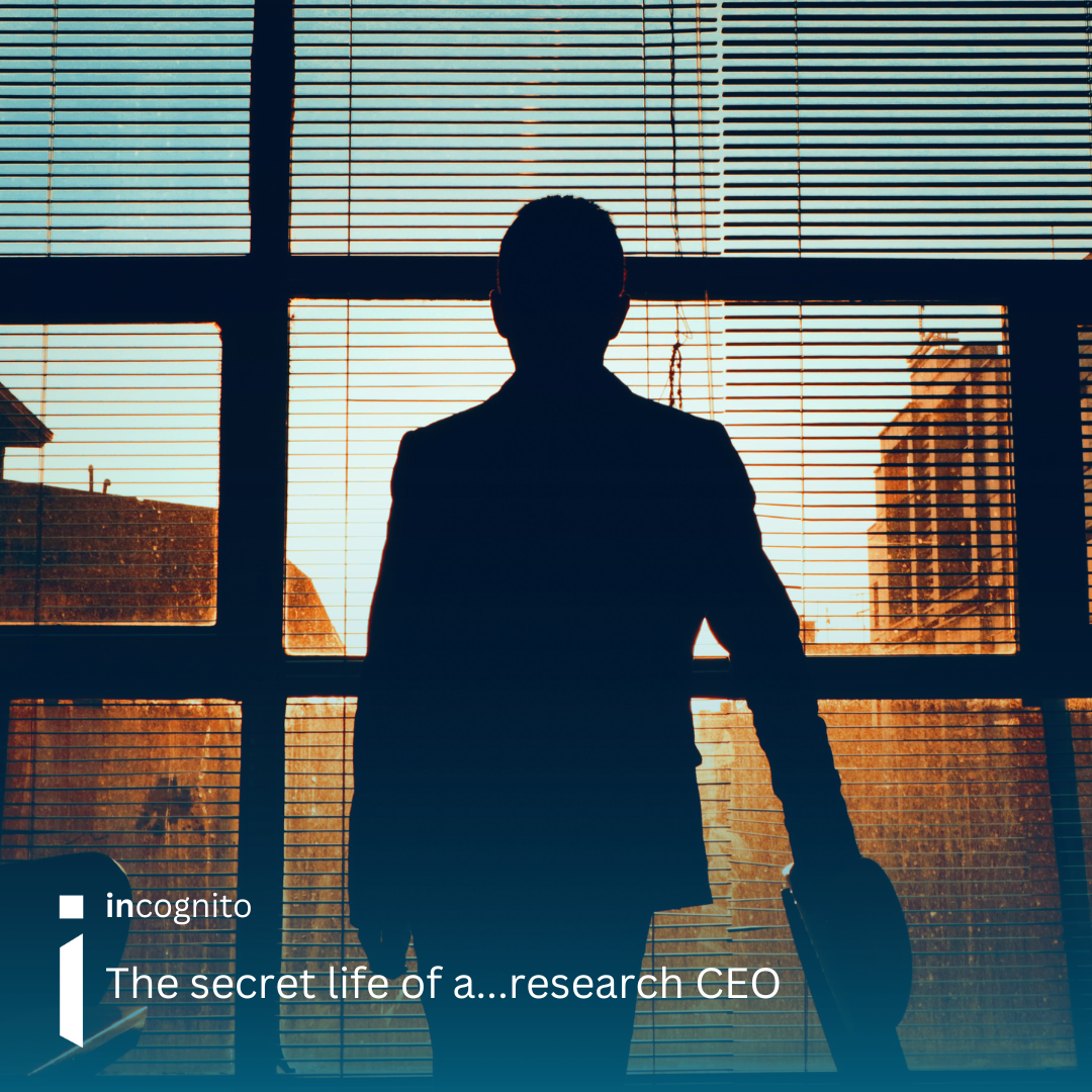 Image of a male, looking away from the camera, mainly in shadow, holding on to a chair on the right hand side, looking out a large office window with horizontal blinds on.