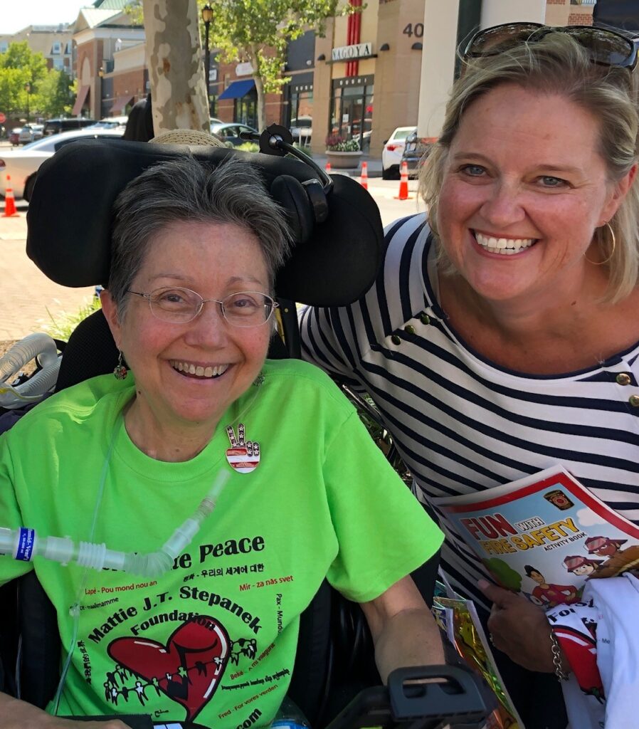 Image of Annie Kennedy on the right wearing a black and white striped top with gold hoop earrings and sunglasses on her head. She is pictured with a lady, on the left, in a wheelchair with a breathing tube. She has short dark hair and a green t-shirt and is smiling into the camera. The background shows shops.
