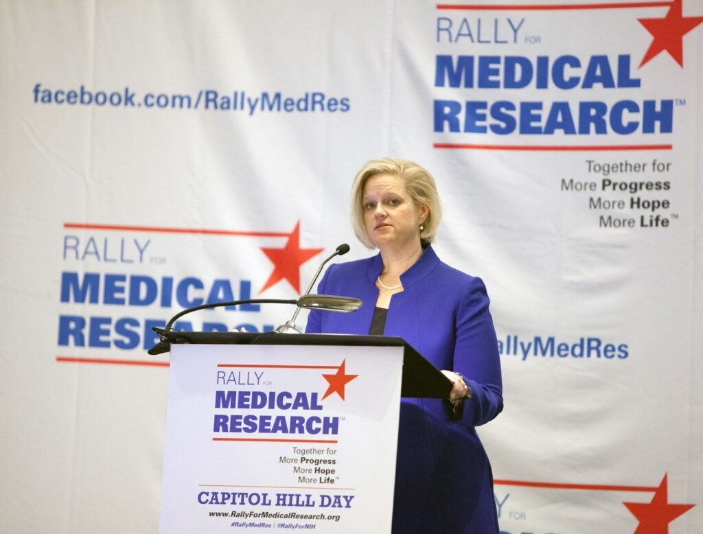 Image of Annie Kennedy standing at a podium with the banner Rally for Medical Research on the podium and behind. She is wearing a purple suit jacket over a black top with a pearl necklace and pearl earrings.