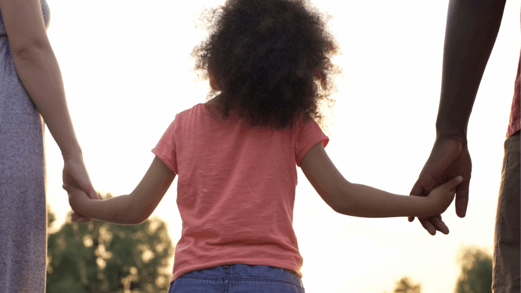 Image of a little girl from the back holding two adults hands either side