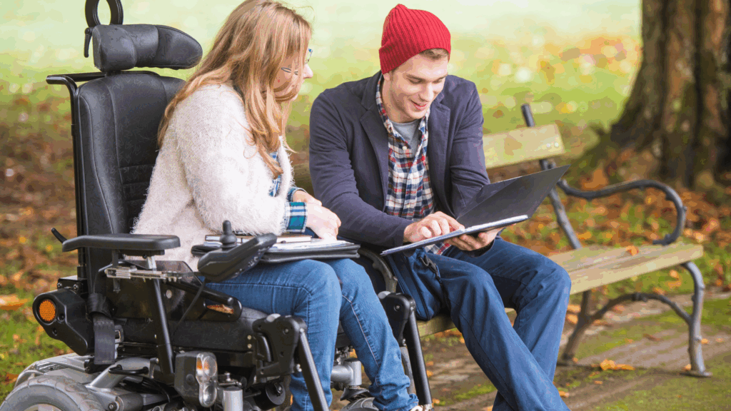 Photo of two young people looking at some work in a folder. The young man is sitting on the bench and the young girl is in a wheelchair.