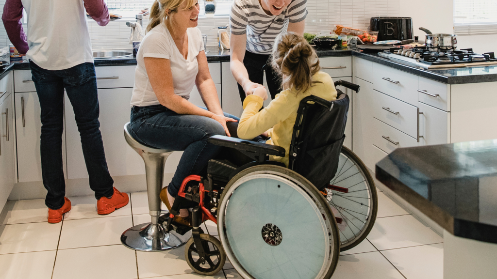 Photo of a girl using her wheelchair, in the kitchen, surrounded by her family