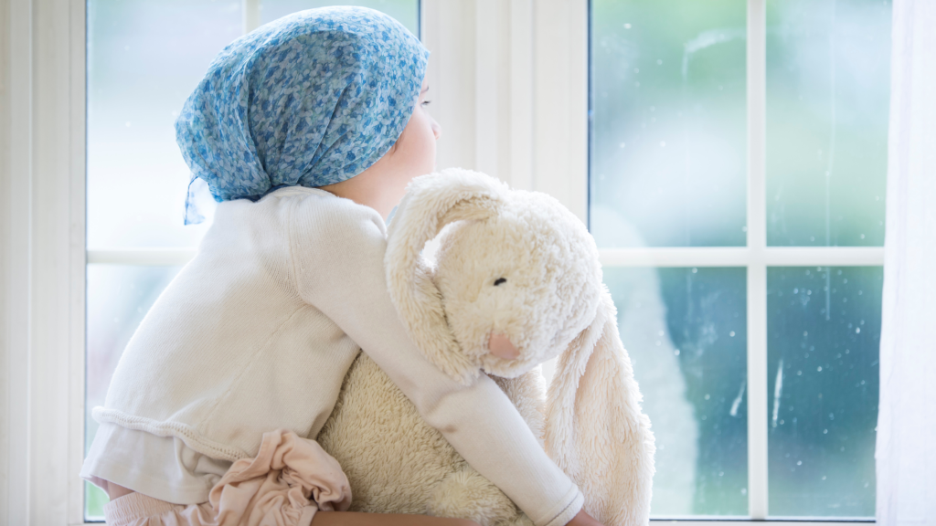 Photo of a child with cancer, hugging a toy rabbit and looking out the window