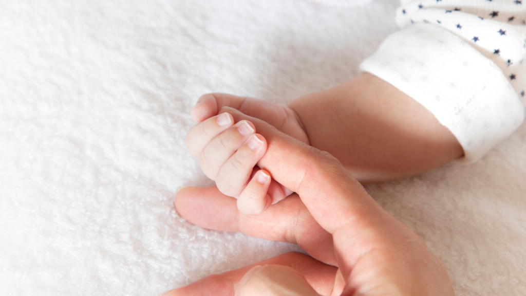 Photo of an adult hand holding a newborn baby's hand.