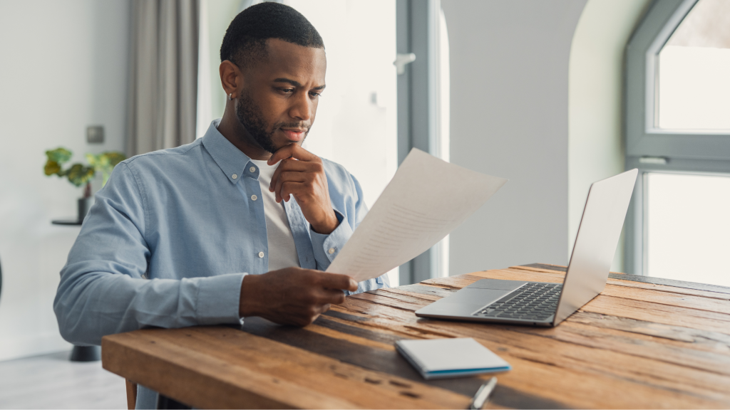 Photo of a man at his laptop reading a document on paper