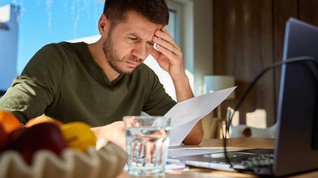 Photo of a man at a laptop, reading a piece of paper, looking confused.