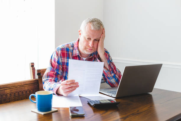 Portrait of a mid adult man in his 30s looking at papers. He has a worried expression and touches his face with his hand while looking at the papers. He is dressed casually in a check patterned shirt.