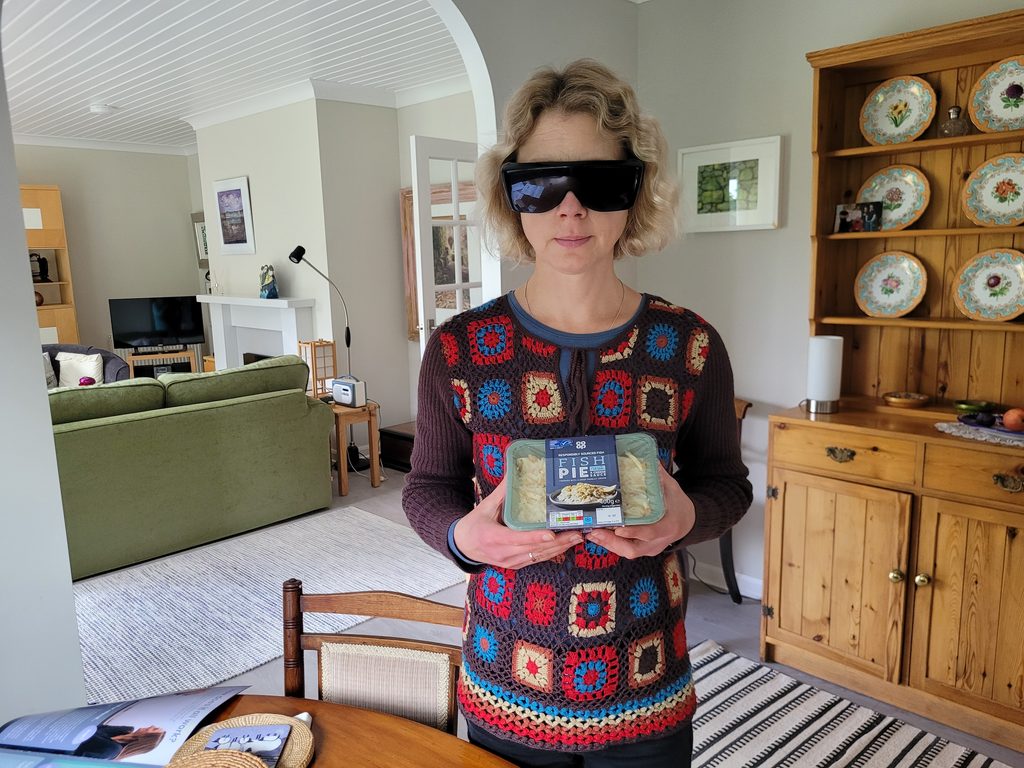 Veronica, a woman wearing a crochet jumper with large sunglasses stands in a kitchen holding a Co-op fish pie. Co-op has braille on its ready meal range.